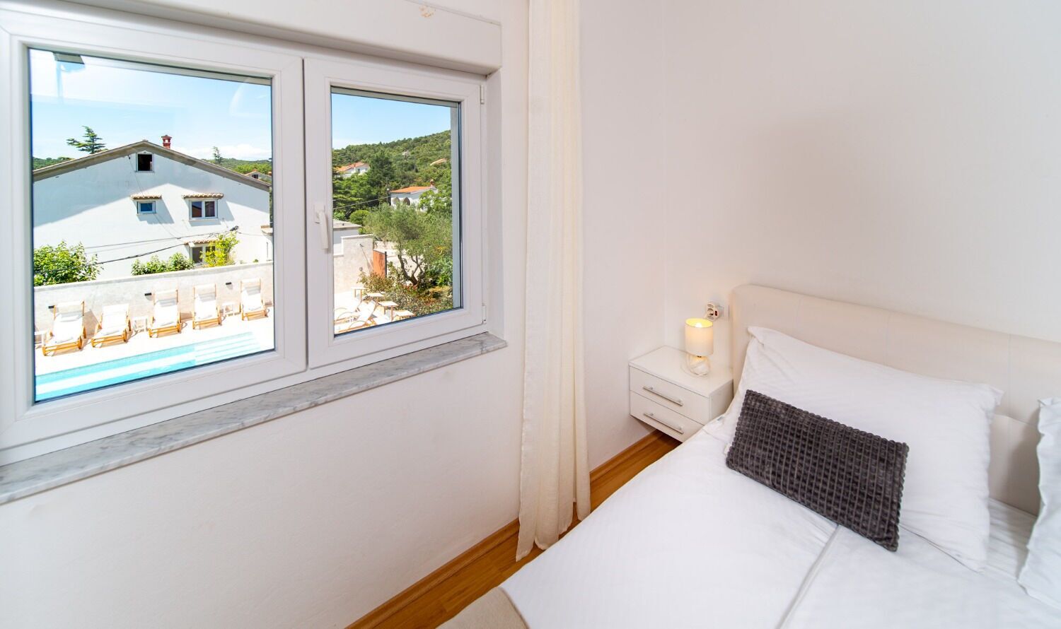 A bedroom with a bed, a nightstand, a lit candle, and a window with a view of buildings and greenery.