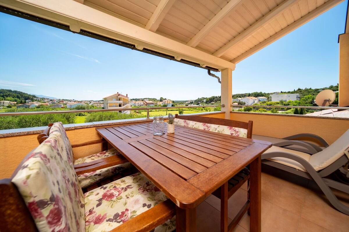 Un espace extérieur aménagé, abrité et doté d’une table en bois et de coussins à motifs floraux, offrant une vue sur un quartier résidentiel avec ses maisons et sa verdure.