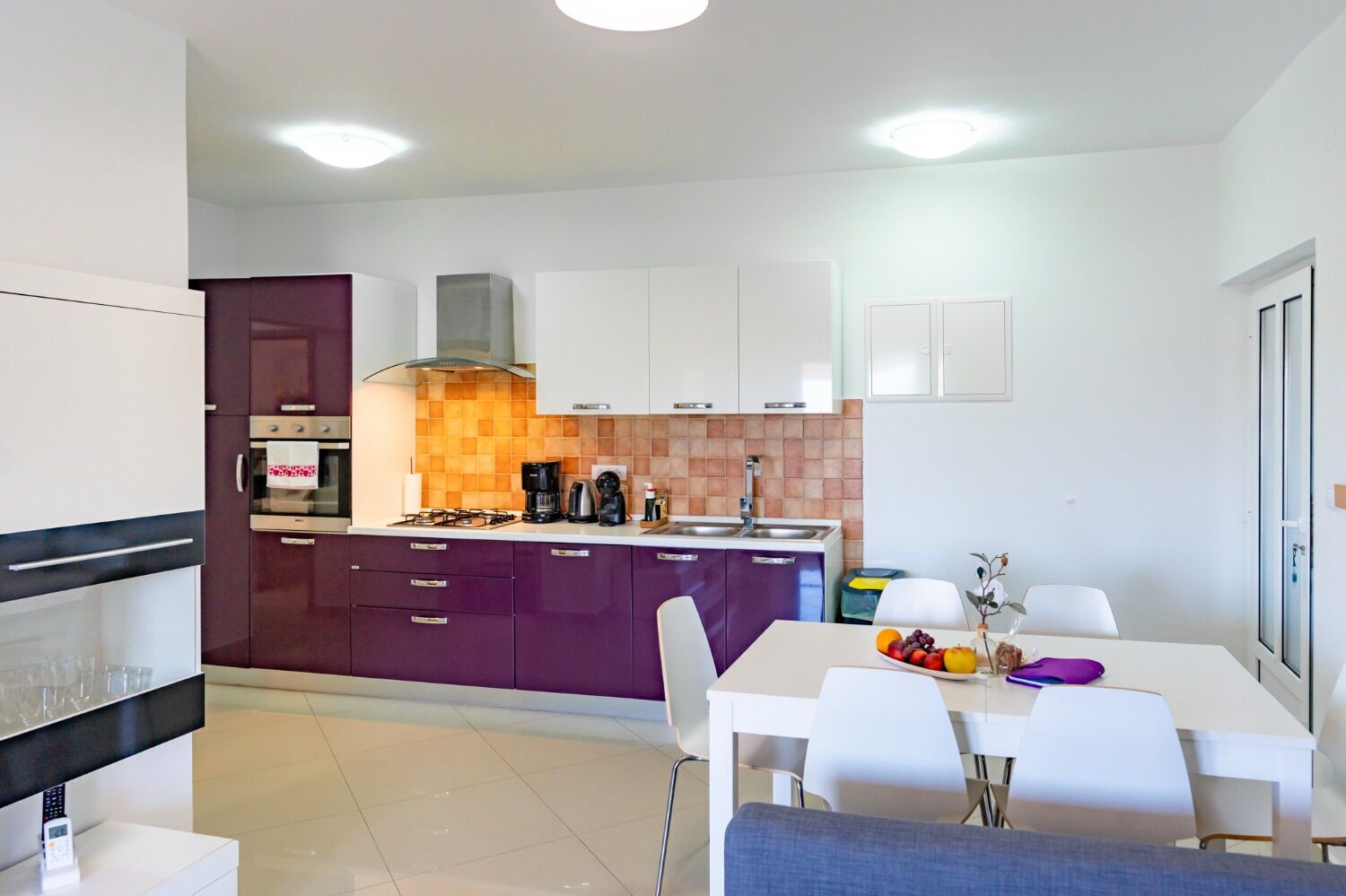 Cocina compacta con electrodomésticos modernos, comedor con mobiliario blanco y vista al exterior a través de una ventana.