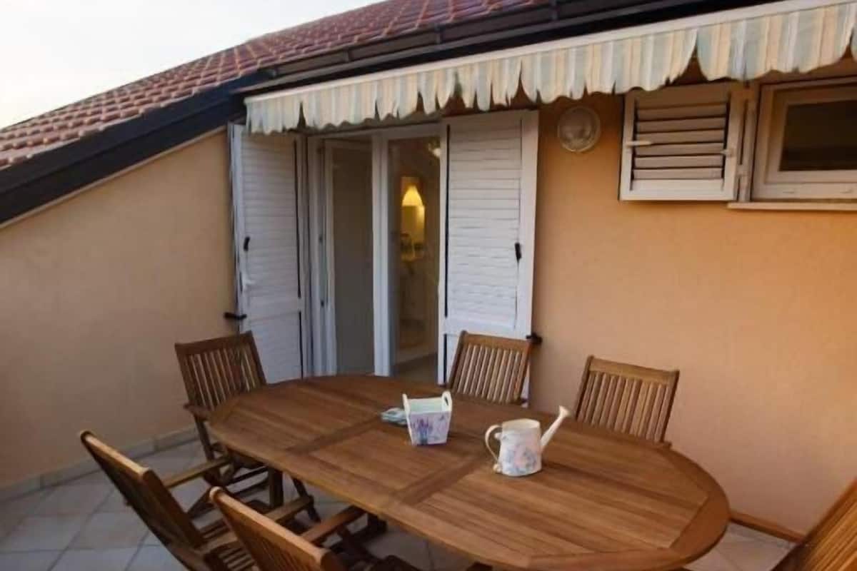 Une table ronde en bois, entourée de six chaises, un pichet blanc et une petite lampe de table, le tout placé sur une terrasse carrelée, surmontée d’un toit recouvert de tuiles rouges et agrémentée de volets blancs.