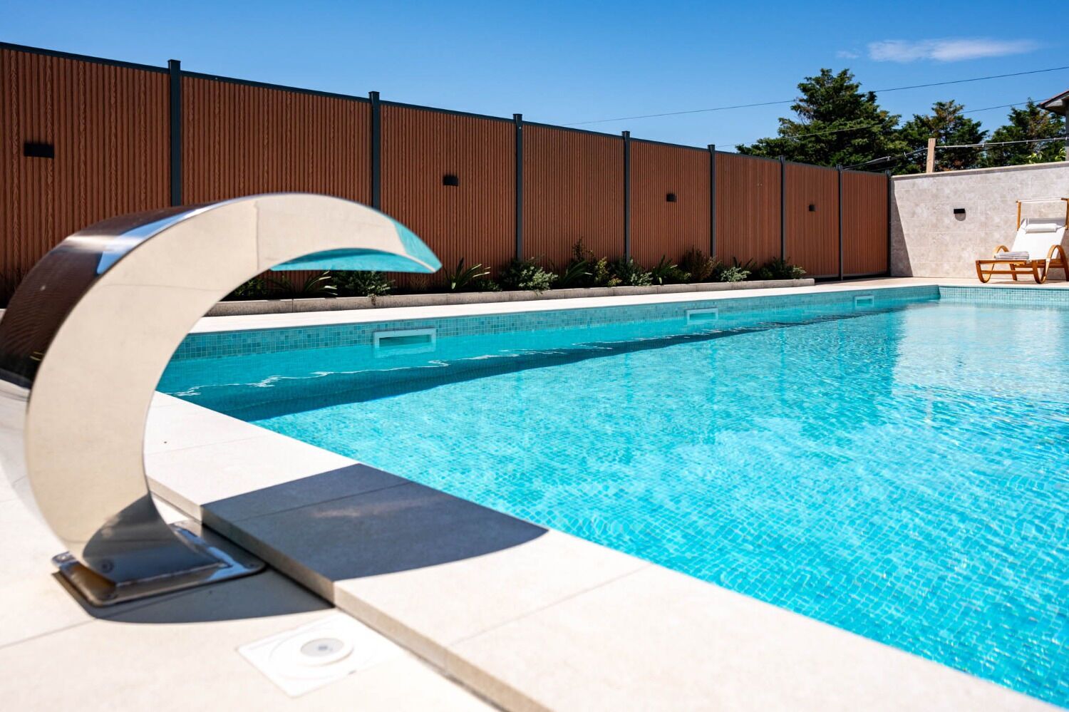 Une piscine moderne à bords incurvés, entourée d’une clôture en bois et d’un mur de pierre.