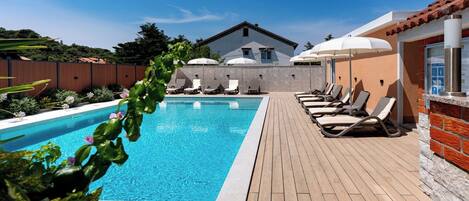 A swimming pool with lounge chairs and umbrellas, surrounded by a wooden deck and a brick wall.