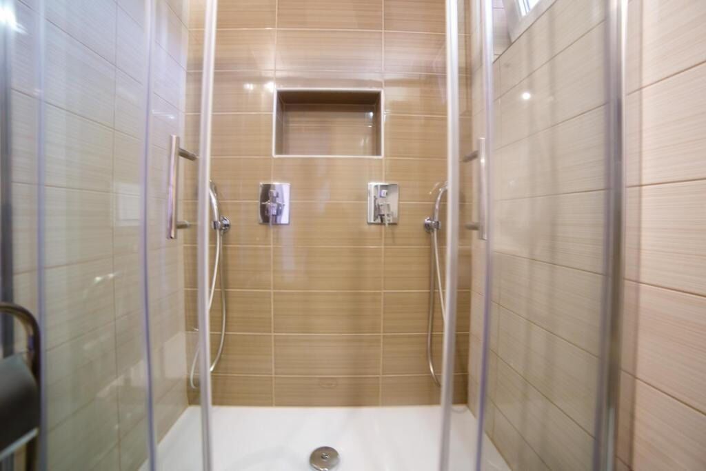 Ducha con cerramiento de vidrio, pared con azulejos, panel de control y un pequeño estante.