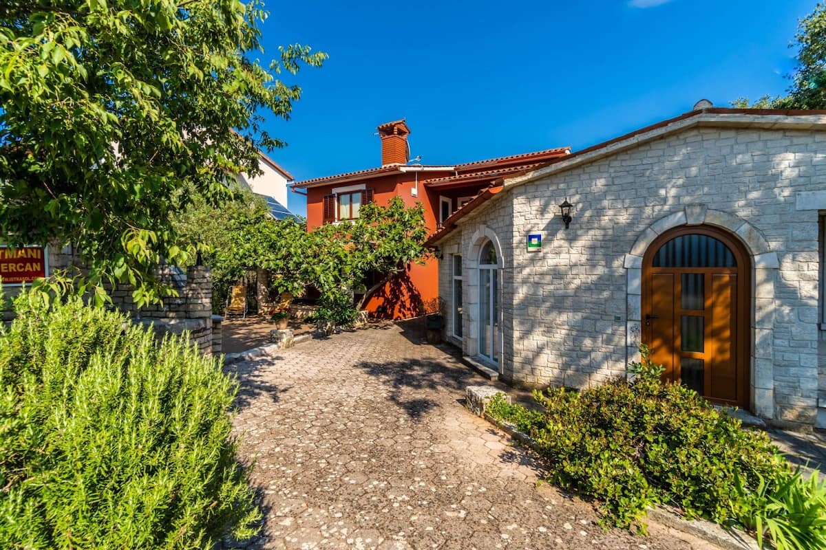Un patio con un sendero adoquinado, un edificio de piedra con una puerta de madera y abundante vegetación.
