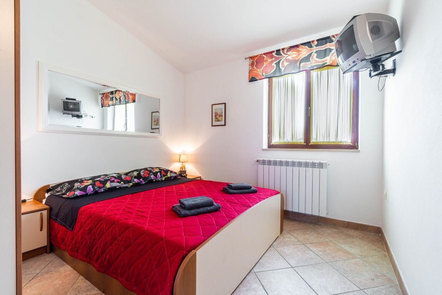 A bedroom with a bed, a nightstand, a window with curtains, a radiator, and a television mounted on the wall.