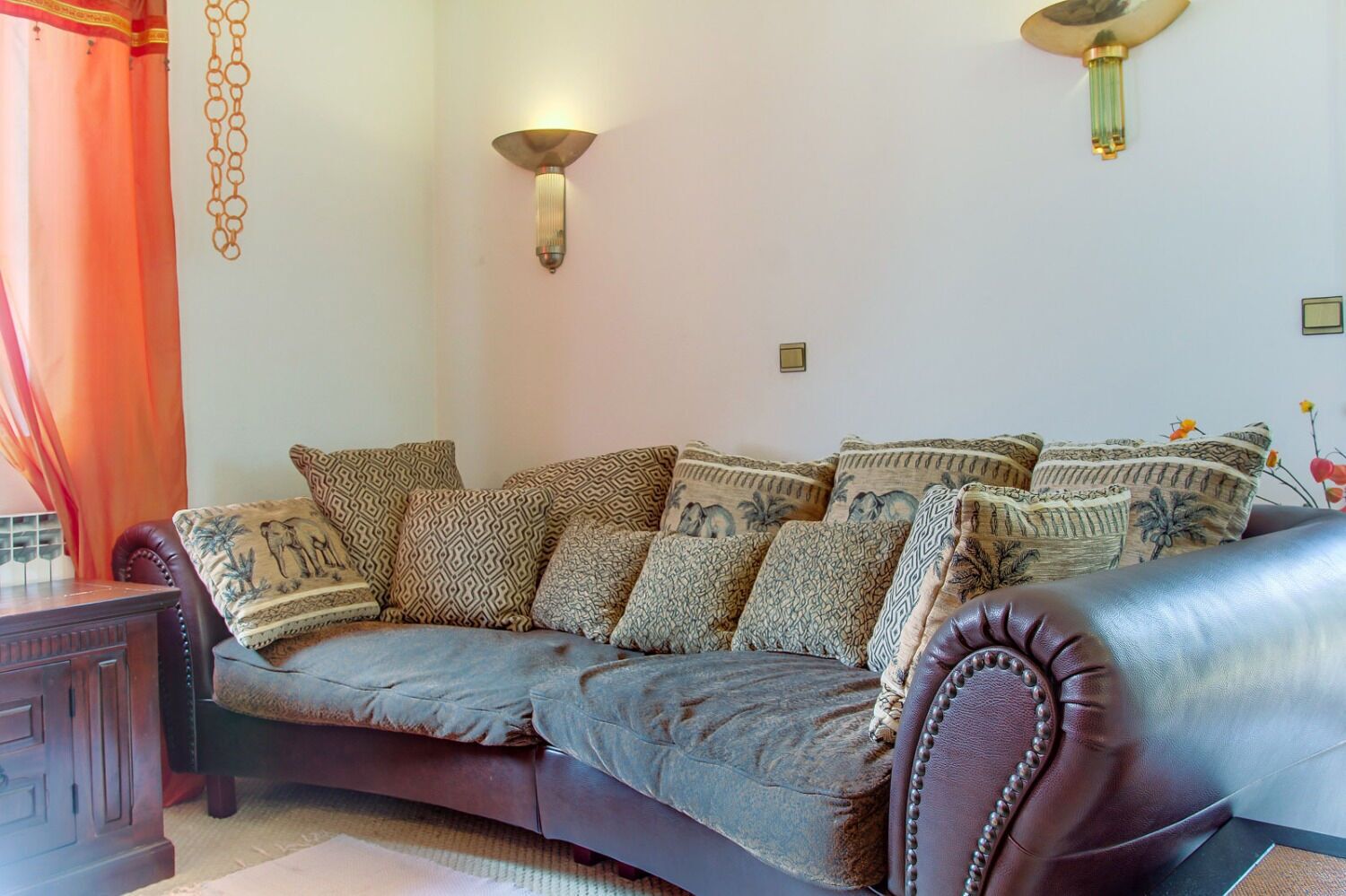 A living room with a dark brown leather sofa, patterned cushions, and two wall-mounted light fixtures.