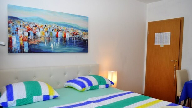 A hotel room with a bed, striped bedding, a painting of a cityscape, a wooden door, and a chair. - Apartments Ivi (Makarska)