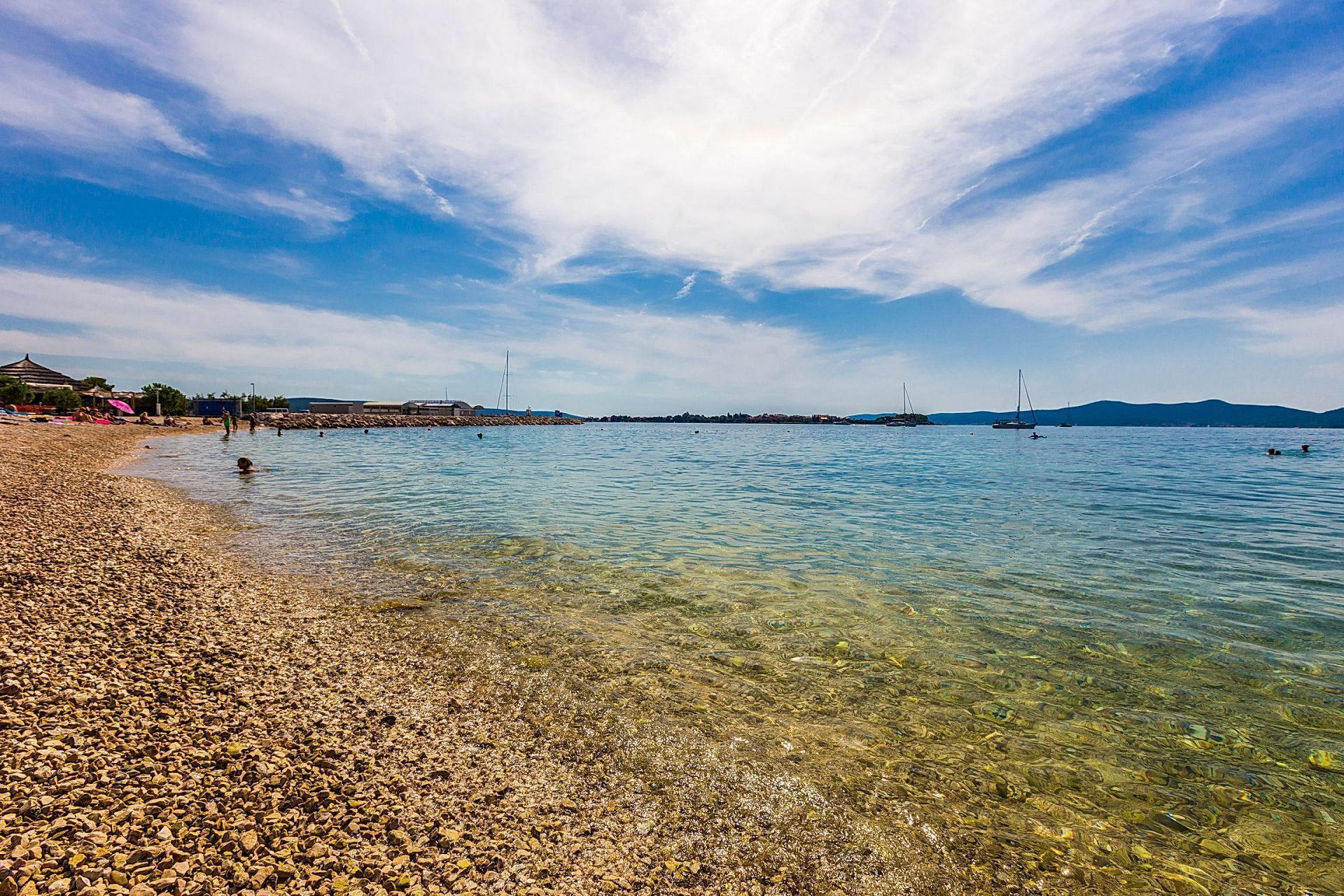 Ein Kiesstrand mit klarem Wasser, Booten und weit entfernten Hügeln.