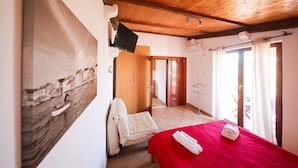 Ein Schlafzimmer mit Holzboden, einem an der Wand befestigten Fernseher, einem Bett mit roter Bettdecke und einem Fenster mit Vorhängen.