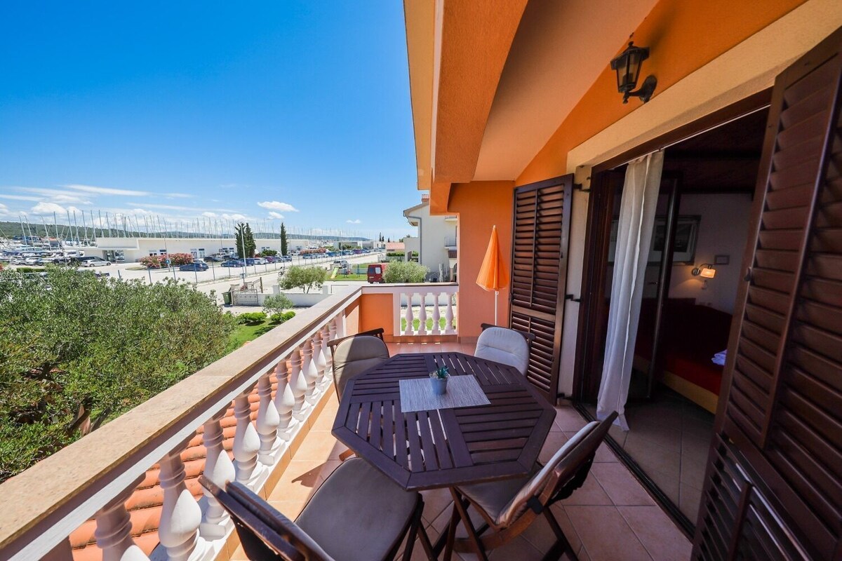 A balcony with a table and chairs, a view of a marina, and a building with orange walls and shutters.