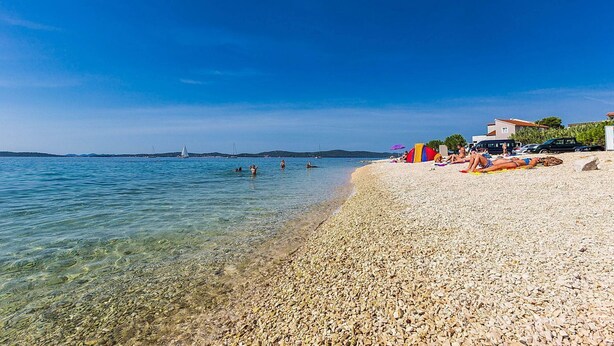 Ein Kiesstrand mit klarem Wasser, einige Badegäste und ein Gebäude mit einem roten Sonnenschirm auf der rechten Seite.