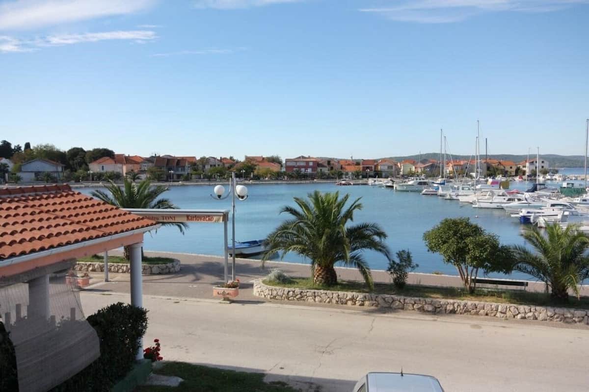 A marina with boats, a sign reading "Nadro Tomi Karovacko", and a building with a red-tiled roof.