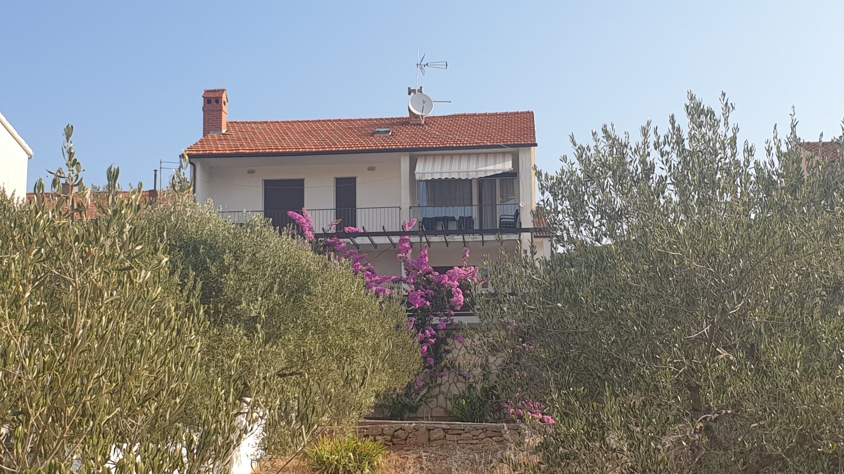 Una casa de dues plantes amb teulada vermella, un balcó amb mobles d'exterior i una paret de pedra al davant.