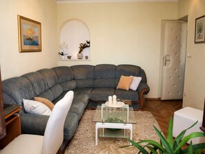 Een moderne woonkamer met een grote grijze modulaire bank, een glazen salontafel en een witte fauteuil. Er is een klein rek met decoratieve objecten en een deur met een gestuikt paneel.