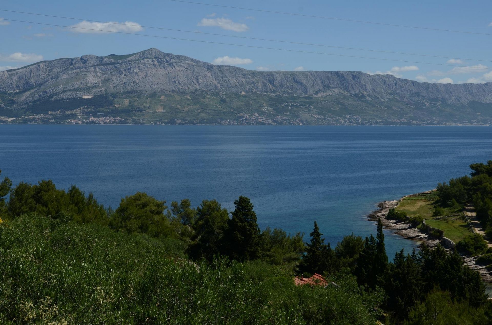 Un paysage côtier avec une chaîne de montagnes en arrière-plan, une mer bleue et limpide, et un petit bâtiment avec un toit rouge situé près du rivage.