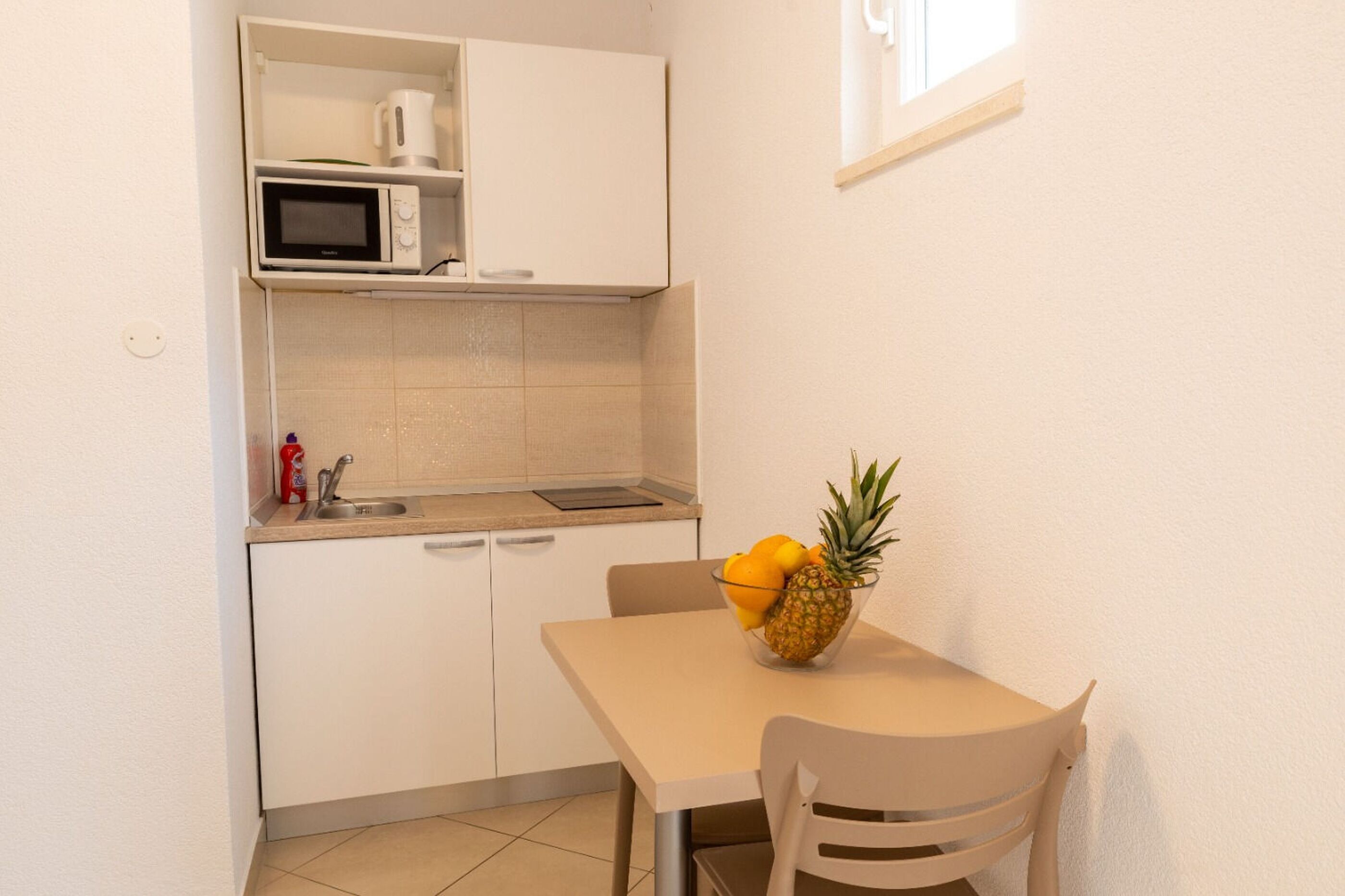 A compact kitchenette with a microwave, sink, and a small cabinet. A table with a fruit bowl and a chair is positioned in front of the kitchenette.