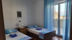Een kamer met twee bedden, een nachtkastje met een lamp, een ingelijst schilderij aan de muur, en een uitzicht door een raam met blauwe gordijnen.