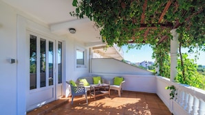 A covered patio with a white railing, a table, two chairs, and a view of the surrounding greenery. - Apartment Pavilion (Rab)
