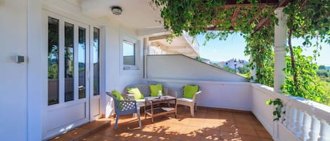 A covered patio with a white railing, a table, two chairs, and a view of the surrounding greenery.