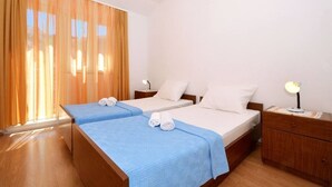 A hotel room with two single beds, a wooden nightstand, a desk with a lamp, and a large window with curtains. - Apartments Sima (Trogir)