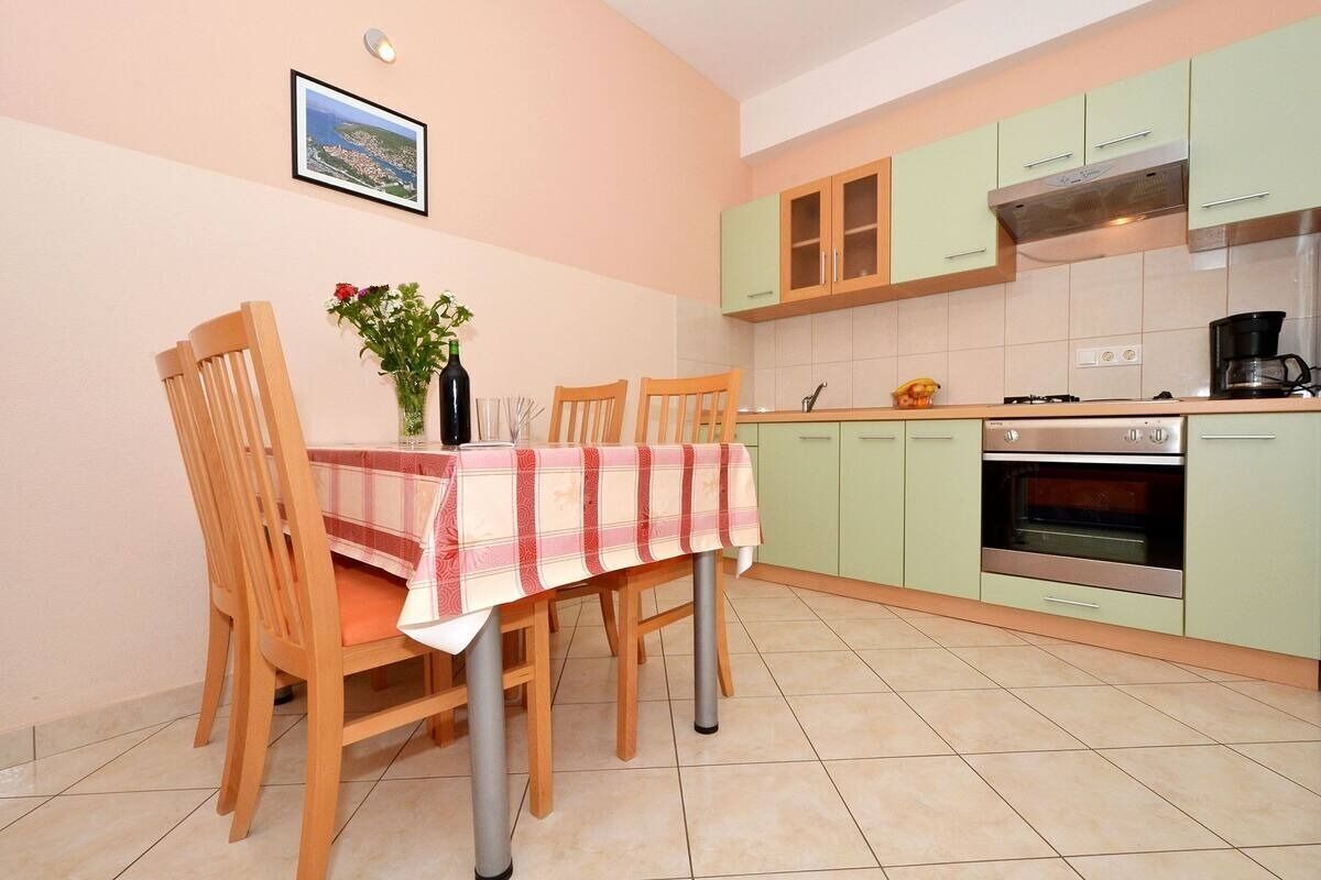 Een keuken met groene kastjes, een eettafel met een geruit tafelkleed en een vaas met bloemen.