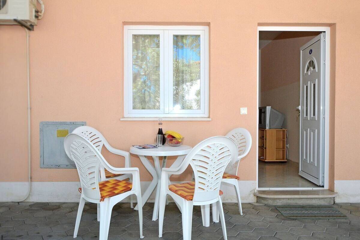 Un petit espace extérieur aménagé avec une table et quatre chaises en plastique blanches, une fenêtre donnant sur des arbres et une porte menant à un espace intérieur.