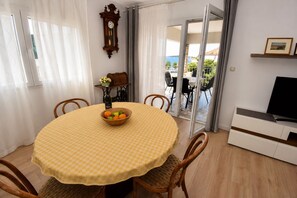 Ένας χώρος τραπεζαρίας με ένα στρογγυλό τραπέζι, καρέκλες, ένα μπολ με φρούτα, ένα ρολόι και μια τηλεόραση τοποθετημένη πάνω σε ένα έπιπλο.