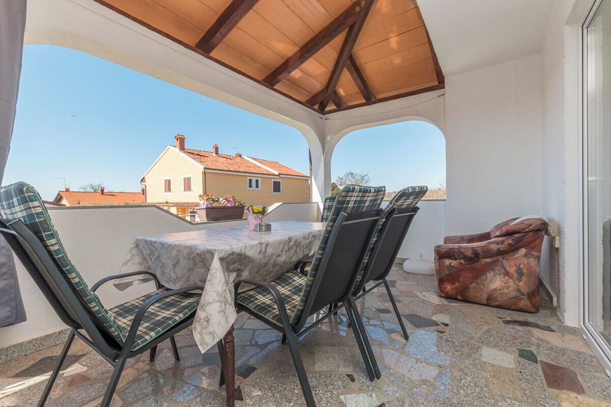 Eine überdachte Terrasse mit Tisch und Stühlen, einer gepolsterten Bank und Blick auf ein Haus im Hintergrund.