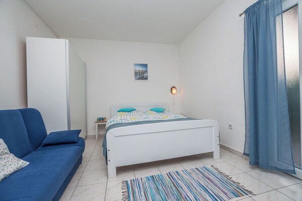 A bedroom with a white bed, a blue sofa, a striped rug, a small bedside table, and a wall-mounted light. - Apartment Neva (Novigrad)