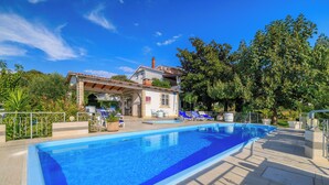 A rectangular swimming pool with a tiled deck, surrounded by lounge chairs and a covered seating area with a pergola. - Apartments Ankica (Rab)