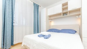 Ein ordentlich bezogenes Bett mit weißer Bettdecke und blauen Kissen, ein weißer Nachttisch mit Lampe sowie ein Fenster mit blauen Vorhängen.