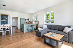 Ampla sala de estar com sofá cinza, mesa de jantar de madeira e área de cozinha com banquetas.