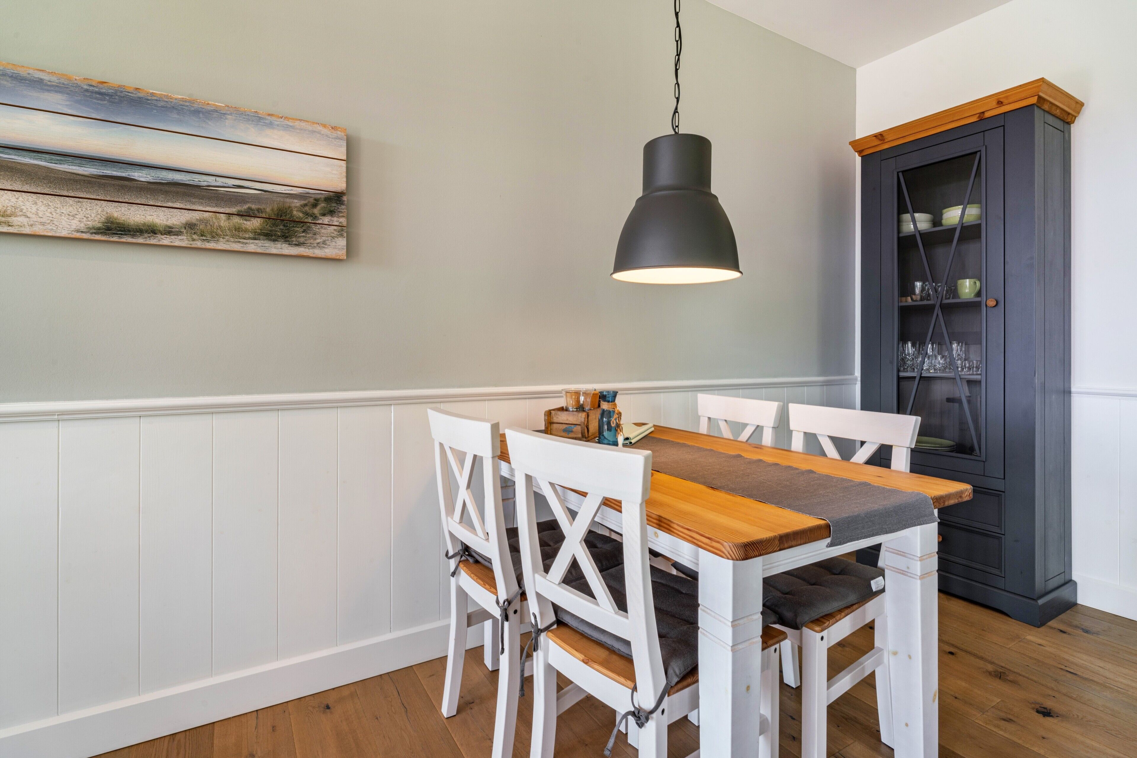 Un coin repas avec une table en bois et des chaises blanches, un luminaire suspendu, un meuble avec des portes en verre et un tableau accroché au mur.