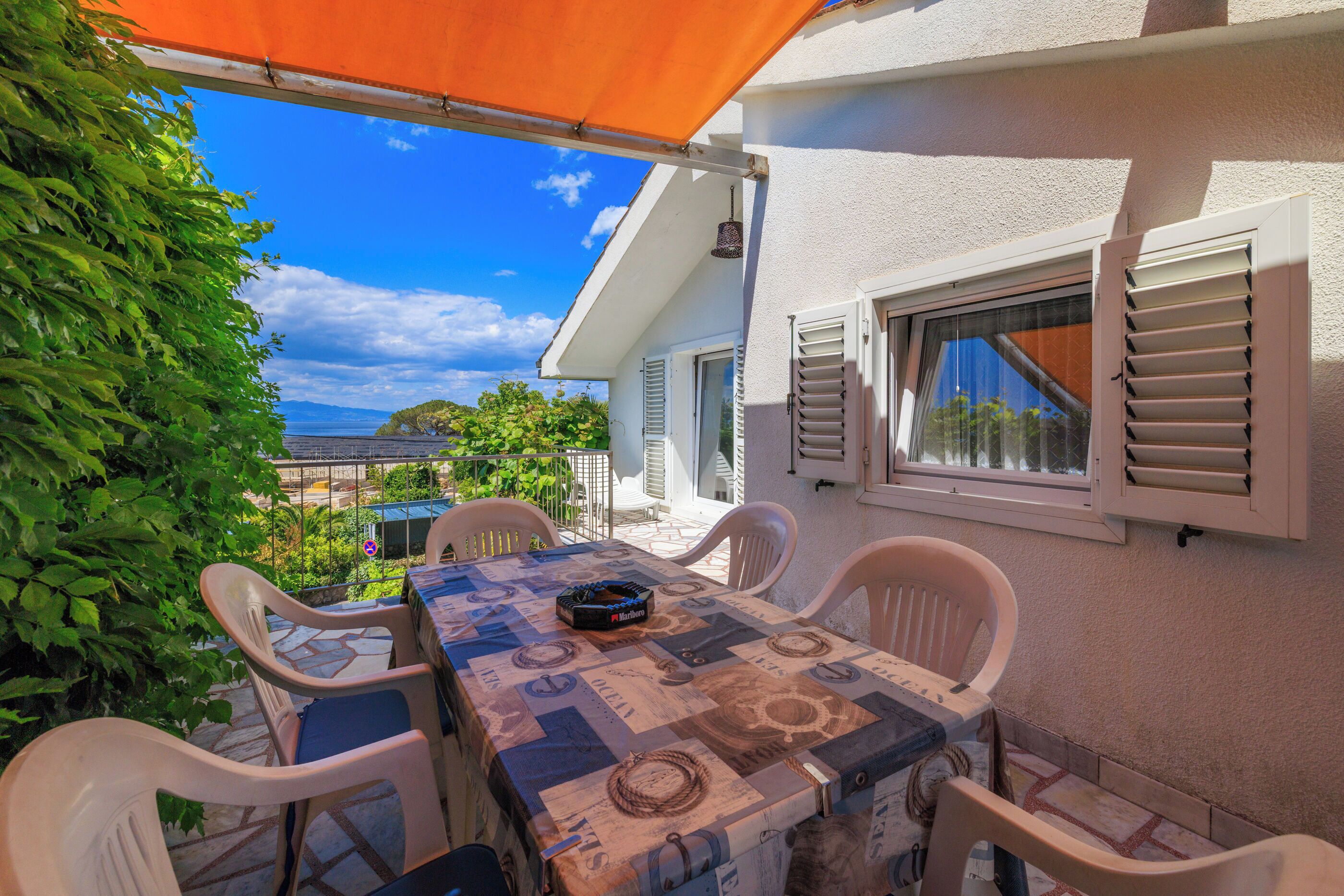 An outdoor patio with a table and chairs, a view of the sea, and a building with shutters.