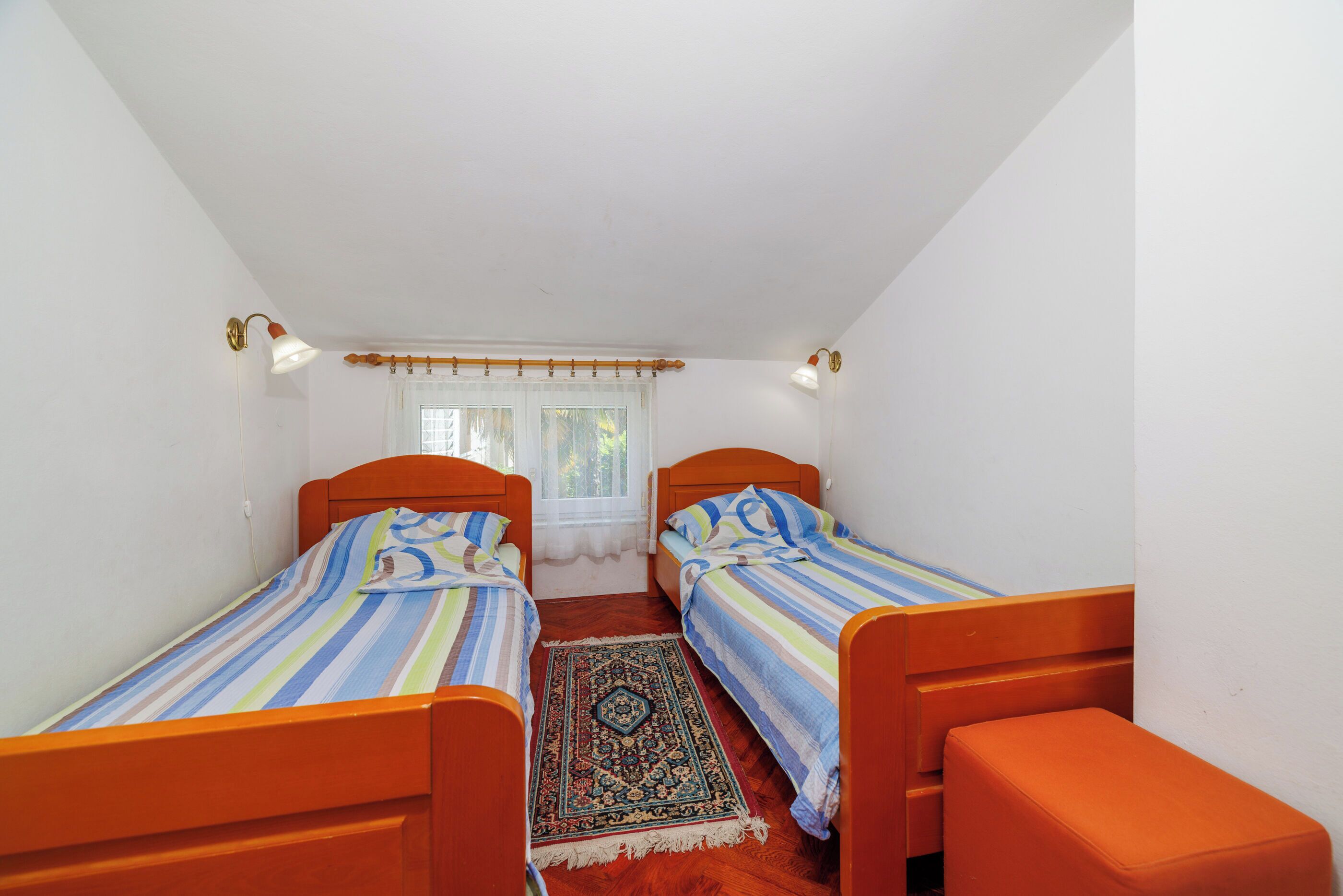 A small, narrow room with two wooden beds, a wooden bench, a patterned rug, and a window with curtains.