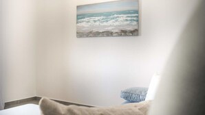 Een kamer met een schilderij van een strandzicht aan de muur, een wit bed en een blauw kussen op de vloer.