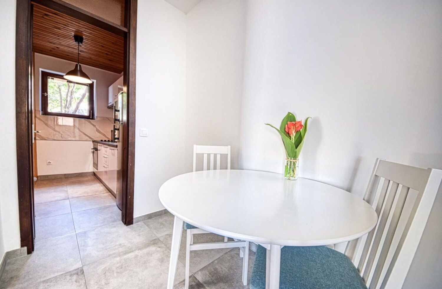 A dining area with a white table and chairs, a vase with flowers, and a kitchen visible through an open door.