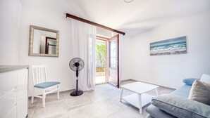 Een moderne woonkamer met een witte bank, een klein wit tafeltje, een staande ventilator en een stoel. Er is een groot raam met witte gordijnen, een schilderij van de zee aan de muur, en een spiegel.