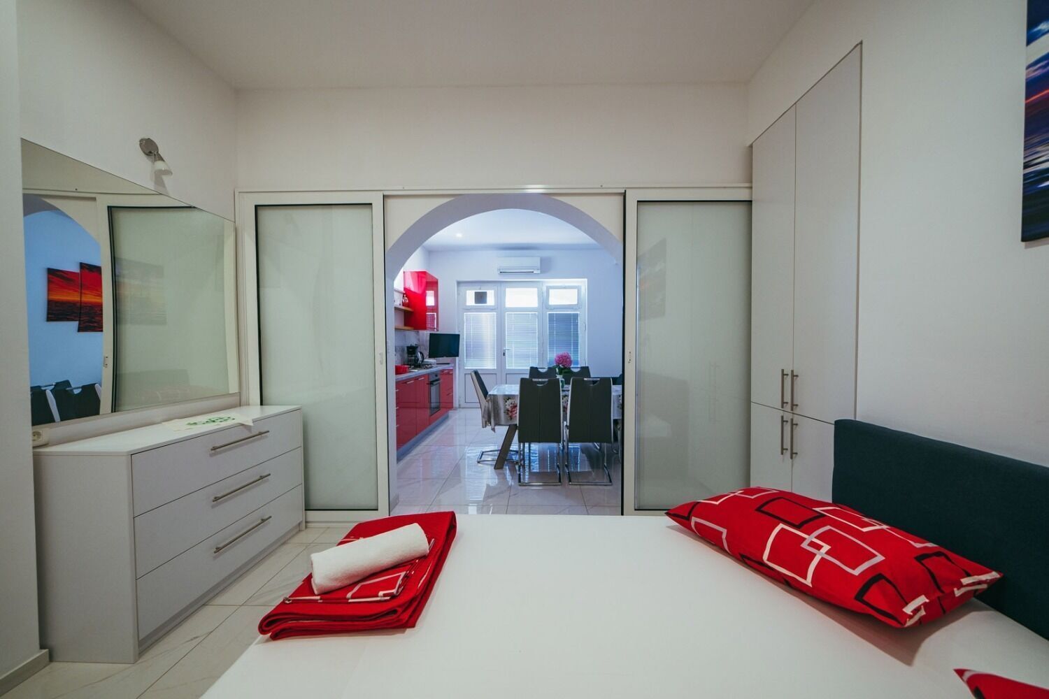 A bedroom with a bed, a dresser, a mirror, and a view into the kitchen.