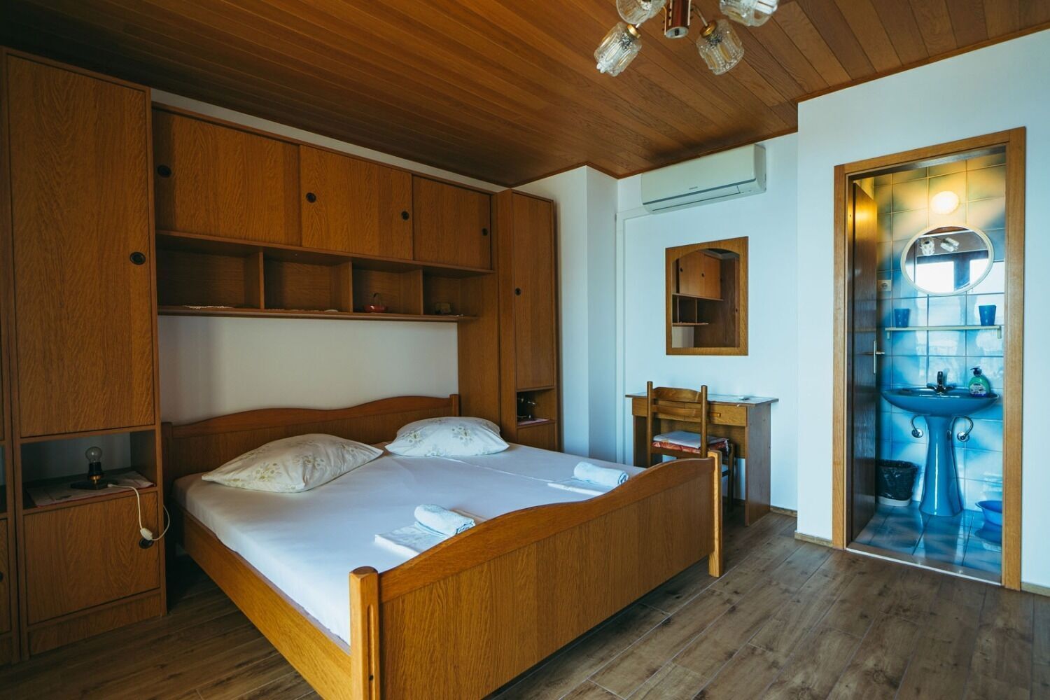 A wooden-framed bed with a nightstand, a desk with a chair, and a bathroom with a sink and mirror.