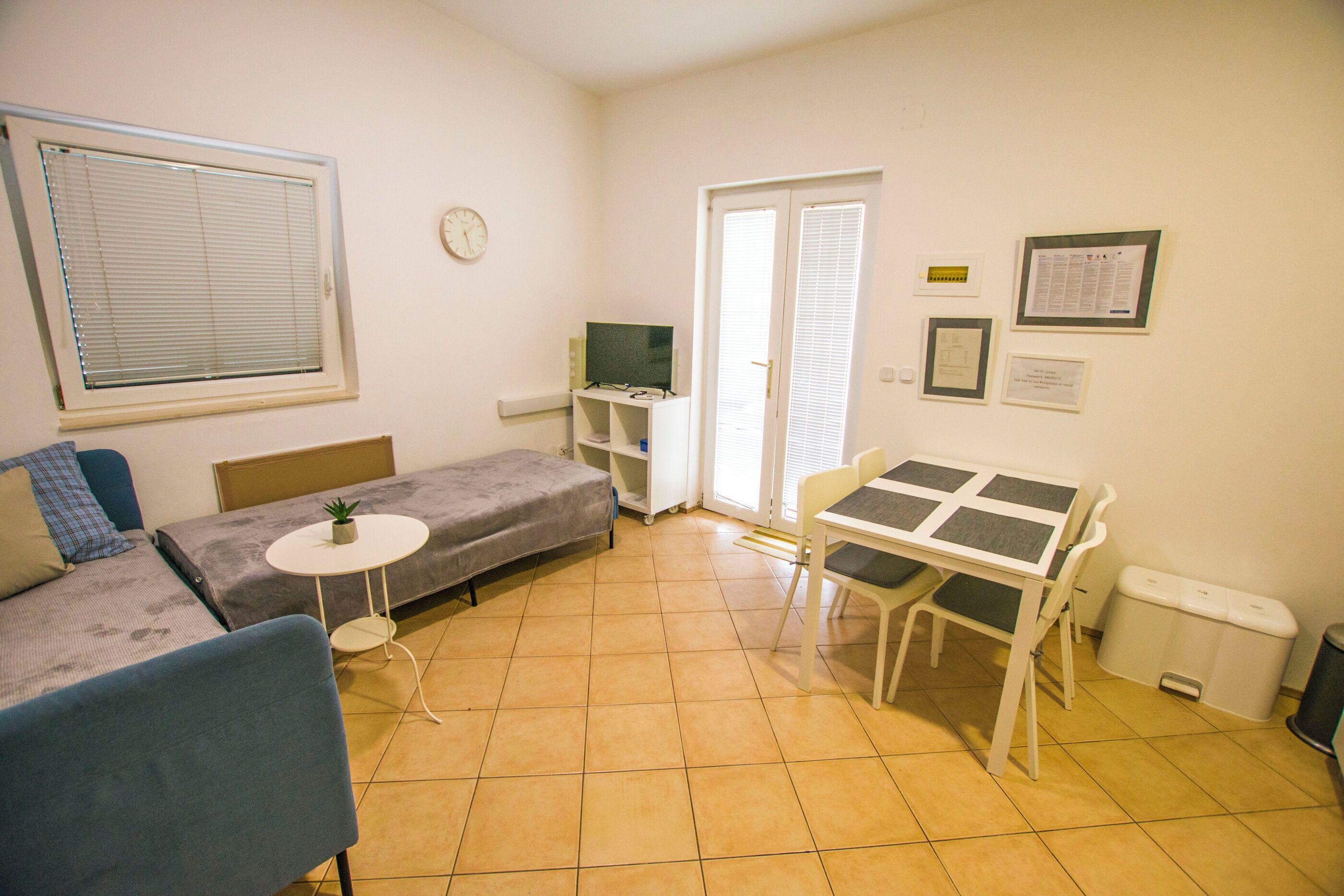 Una habitación pequeña y bien iluminada con un sofá, una mesita, una silla, un televisor y una ventana con persianas.