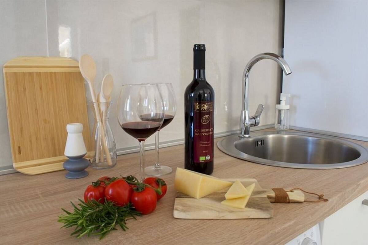 A kitchen counter with a bottle of wine, glasses, tomatoes, cheese, and a cutting board.