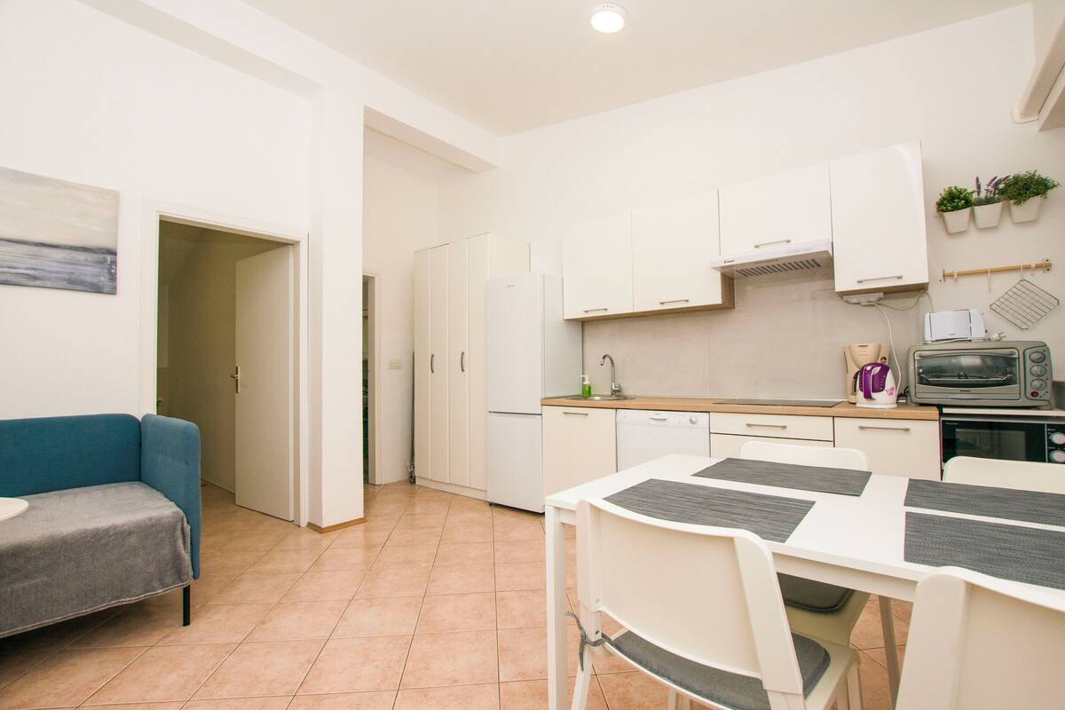 A compact kitchen with white cabinets, a microwave, and a toaster. There is a dining table with chairs and a sofa in the adjacent living area.