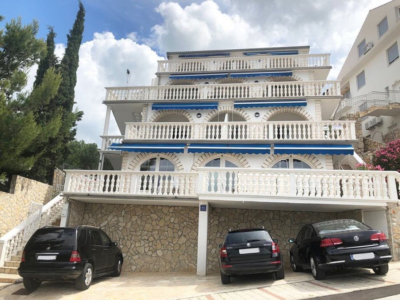 A multi-story building with a stone facade, balconies with white railings, and three parked cars in front.