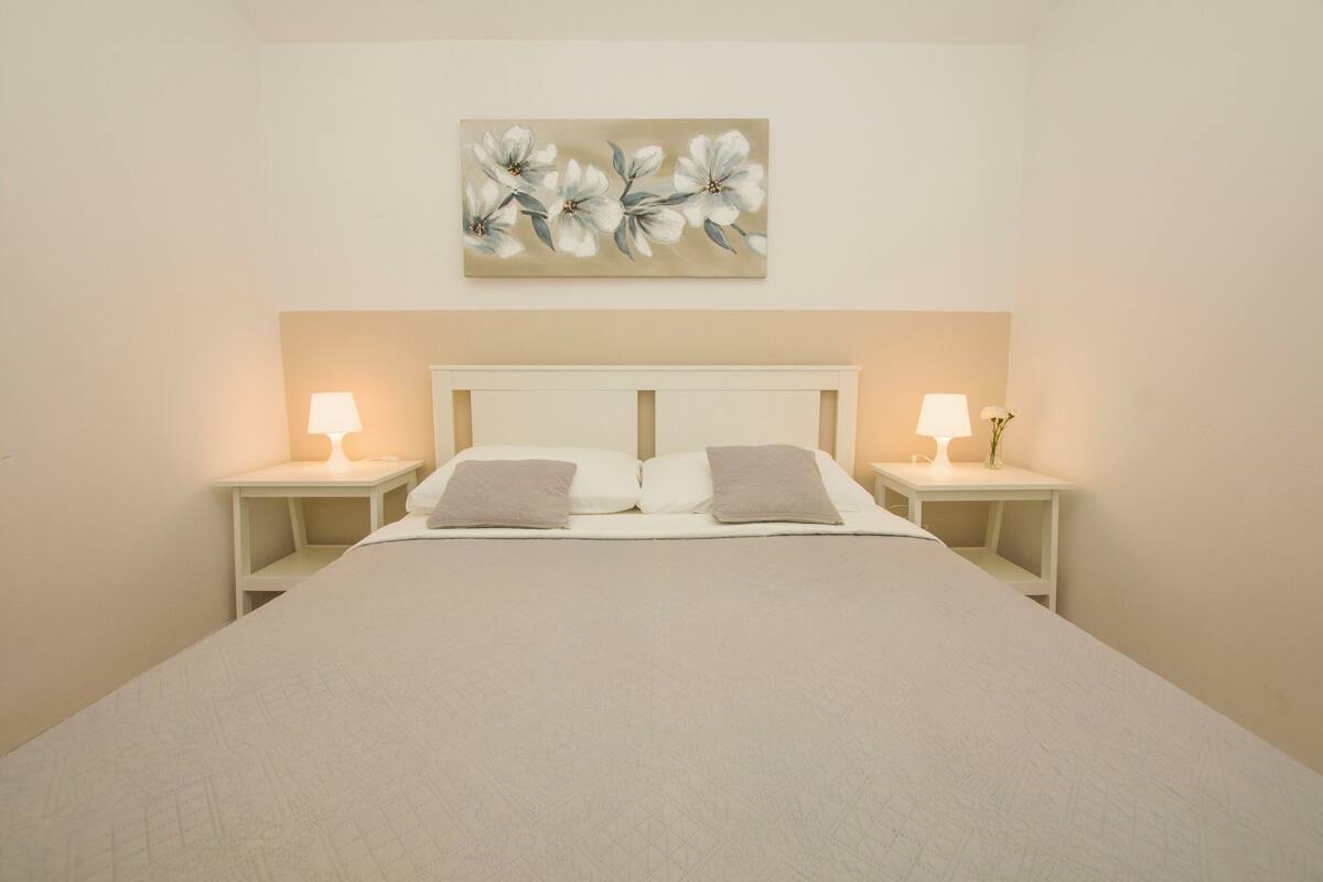 A neatly made bed with a white headboard, two bedside tables with lamps, and a floral painting above the headboard.