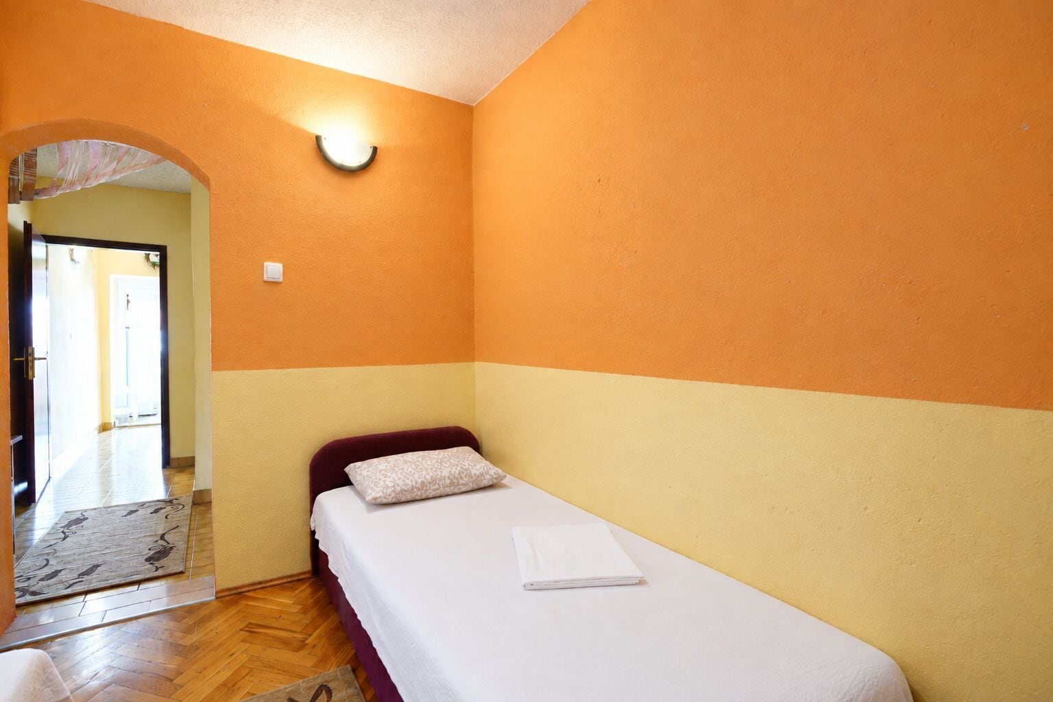 Un lit simple avec un matelas blanc et un oreiller à motifs, dans une pièce aux murs orange et au plancher en bois.