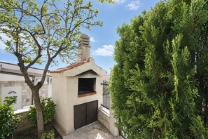 A small outdoor structure with a chimney, possibly a barbecue area, surrounded by trees and a building in the background. - Apartment Lile (Omis)