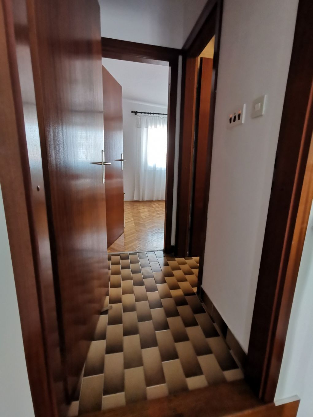 A hallway with a checkered floor pattern, wooden doors, and a view into a room with a curtain and a window.