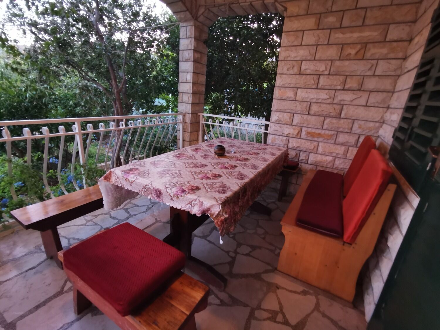 An outdoor patio with a table and benches, surrounded by stone flooring and a white fence.