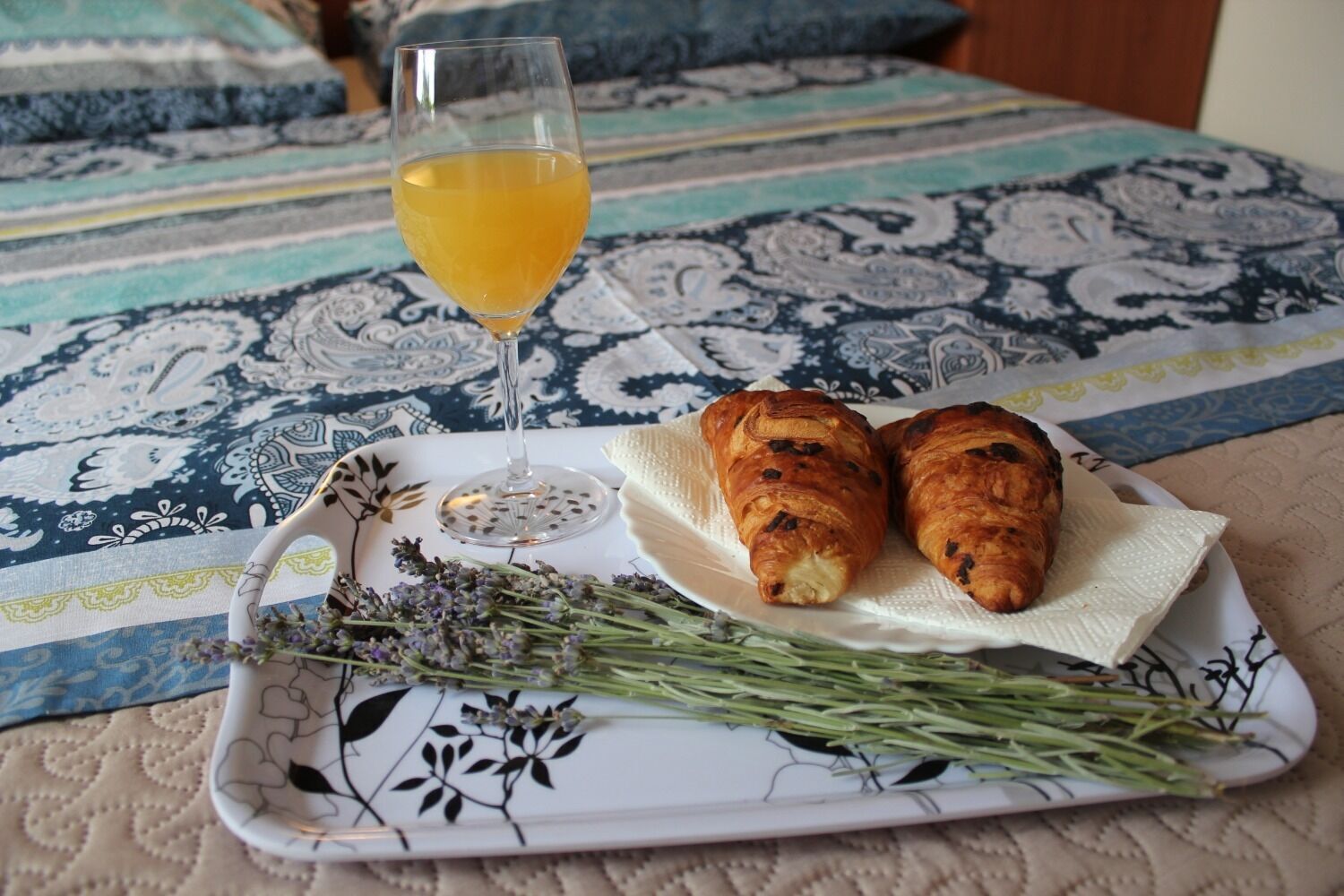 En bakke med to croissanter, et glas appelsinjuice og en rosmarin kvist på en mønstret dug.